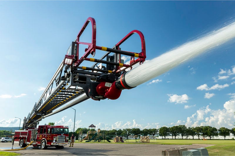 The ladder of a quint fire truck is extended spraying water in the foreground while the red truck is parked in the background with a blue sunny sky. 