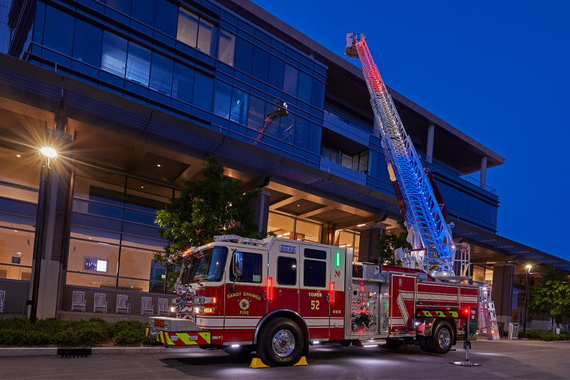 A red and white quint fire truck is parked at dusk with its ladder raised at a 3-story building. The ladder is lit red, white and blue and the fire truck emergency lights are turned on.