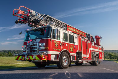 A Pierce ascendant aerial ladder apparatus drives along hilly, rural terrain.