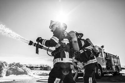 Two firefighters hold a spray nozzle shooting water from a pumper truck at the scene of a fire.