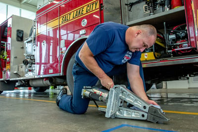 A firefighter works with a tool on the ground next to a parked fire truck inside a station house. 