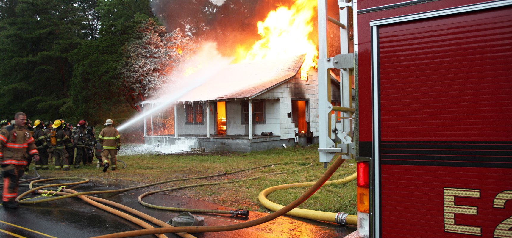 A live fire burning house with fire fighters and a Pierce fire apparatus on the scene using a fire-fighting foam system to put out the fire.