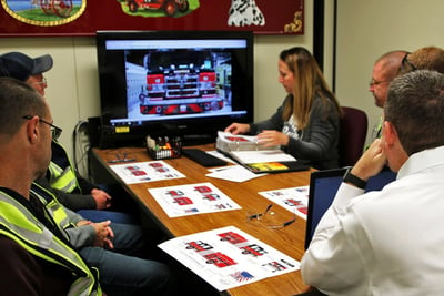 Pierce Manufacturing team members and customers sit around a table reviewing fire truck drawings and images on paper and on a screen. 