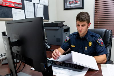 A firefighter reviews the details of a fire truck order contract in his office behind a desk and monitor. 