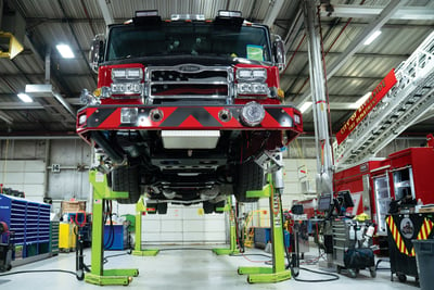 A fire truck is elevated on a lift inside a mechanic facility as is undergoes fleet maintenance.