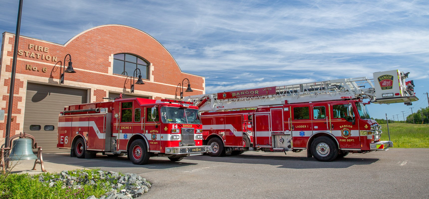Two red and white fire trucks are parked outside their fire department bays with a bright blue sky in the background. 
