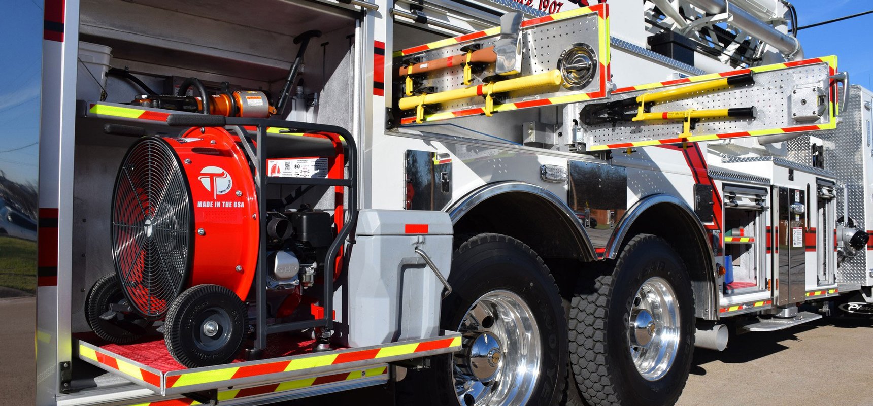 A Pierce aerial apparatus is parked showing the driver side storage options, including a pull-out tray with a fan, peg swing boards with on-scene equipment and tools and storage compartments. 