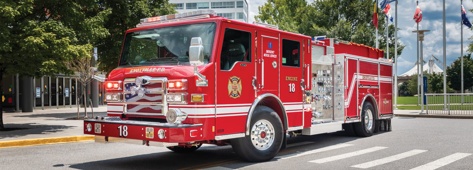 A red type 1 fire truck is parked in an urban setting with flags in the background.