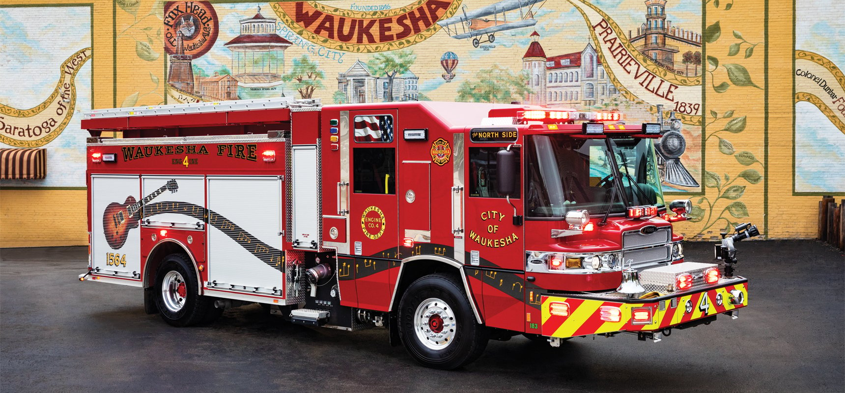 Pierce fire truck, with Les Paul guitar graphics, positioned in front of a historical and community themed mural downtown Waukesha, WI.