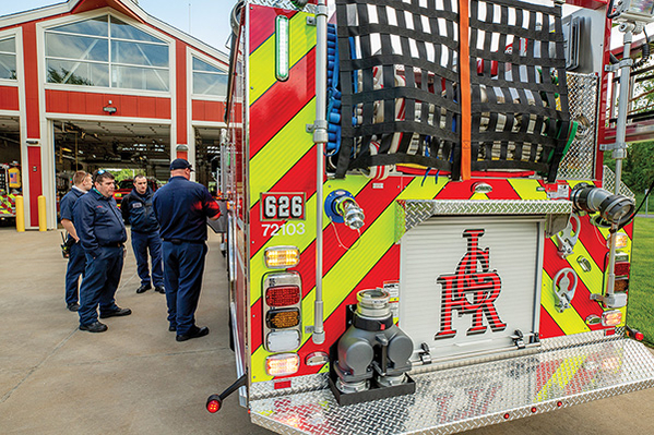 Rear hose bed of pumper fire truck with firefighters standing next to in front of fire station