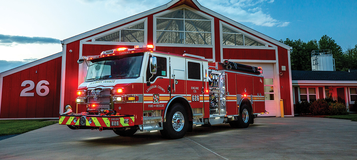 Pumper fire truck with emergency lights on parked in front of red fire station at dusk