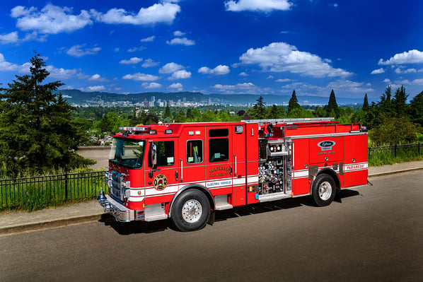 The driver's side of a Pierce Electric Fire Truck parked in front of green trees and mountains on a sunny day with clouds in the sky.