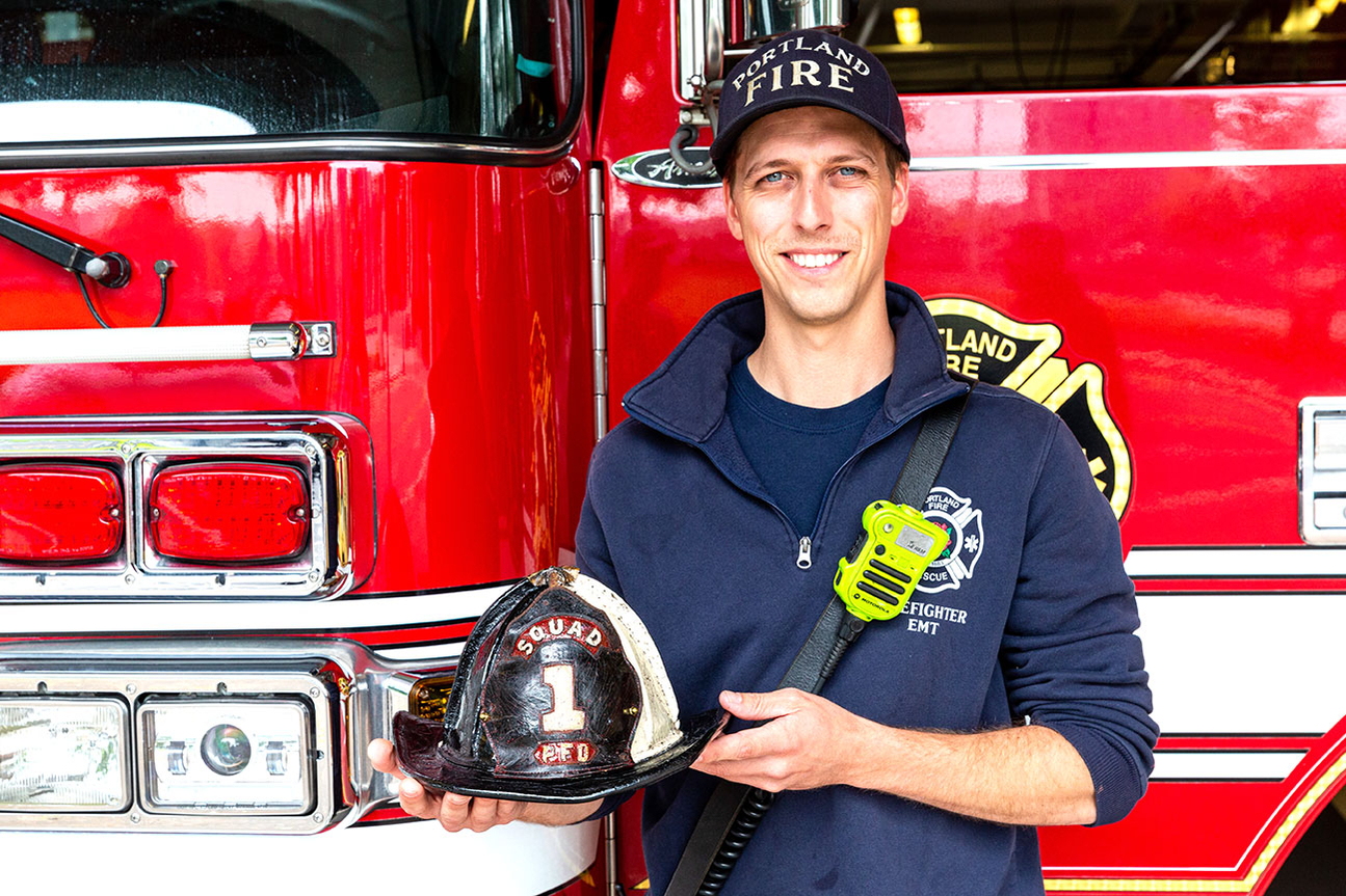 Portland Oregon firefighter standing and smiling in front of fire engine parked in station holding fire helmet