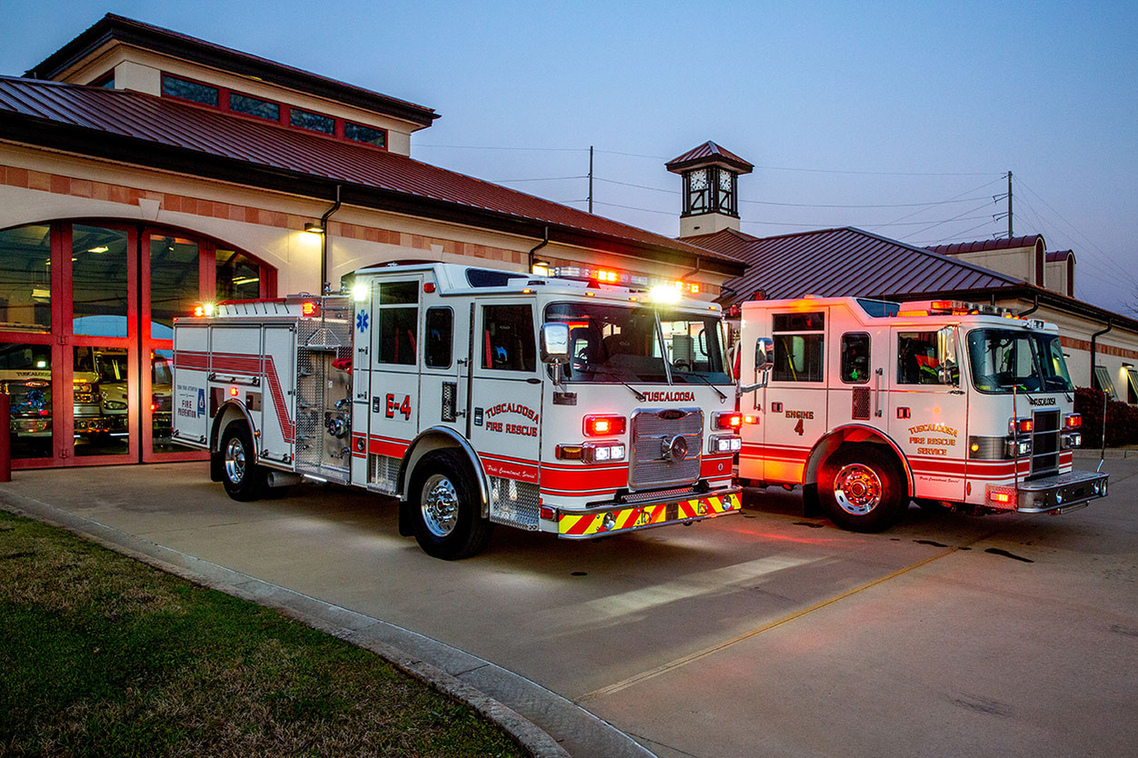 Two pumper fire trucks parked on apron of fire house at dusk with lights on