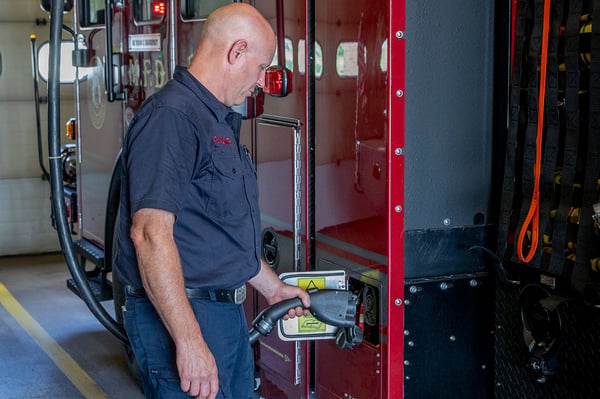 A firefighter at a fire station plugging in the charging cord to a Pierce Electric Fire Truck on the driver's side.