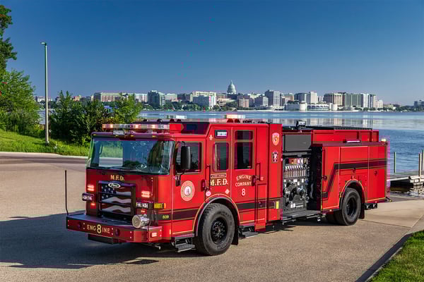 The drivers side of an Enforcer Volterra Electric Fire Truck in front of a a lake and city skyline on a sunny day.