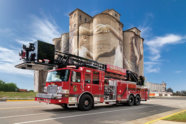 The front drivers side of an Enforcer 100’ Heavy-Duty Steel Aerial Platform driving on a road in front of a historic building.
