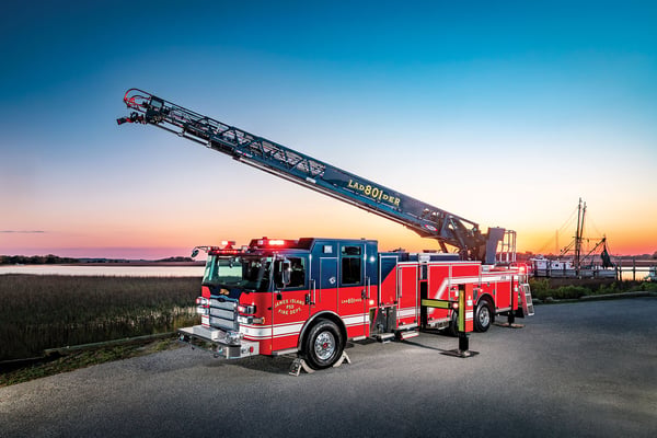 The front drivers side of an Enforcer PUC 107 Ascendant Aerial Ladder parked in front of the ocean and a sunrise with the aerial up.