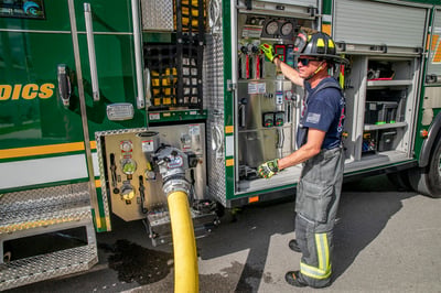 A firefighter in turnout gear operting the pump of a PUC Pumper fire truck and a charged fire hose coming from the discharge.