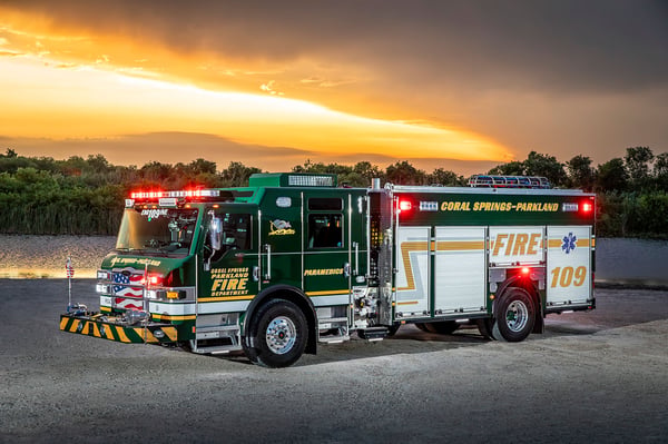 The front drivers side of an Impel PUC Pumper parked on gravey by water and a marsh area with a sunset behind.