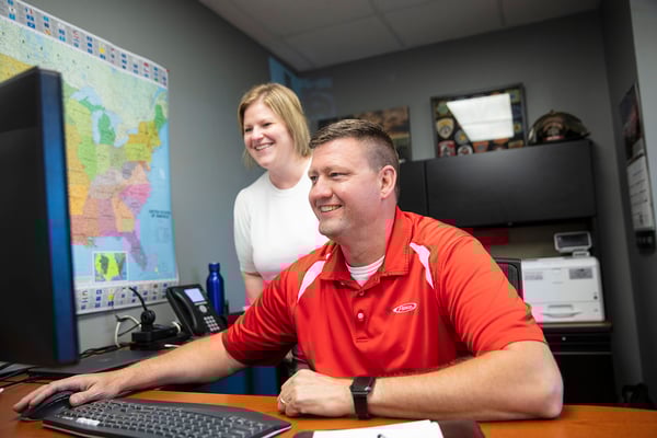 Two office team members looking at a computer screen with a map of the United States on the wall.