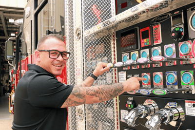 A man in a black polo shirt with tattoos on his arm and black glasses points out the gauges on a fire truck.