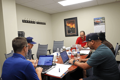 A group of men sit around a conference room table with papers and computers in front of them.