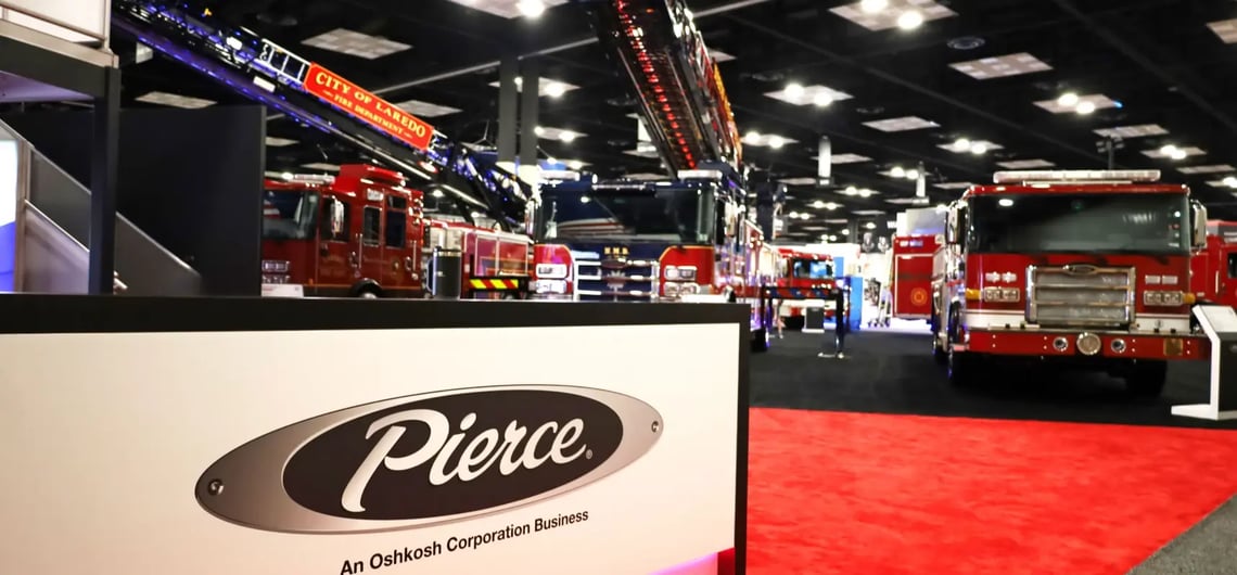 Pierce fire trucks exhibited at a trade show with a Pierce brand booth sign in the foreground.