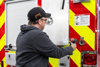 A team member putting the rear compartment door on a fire truck with a drill in a modern manufacturing facility.