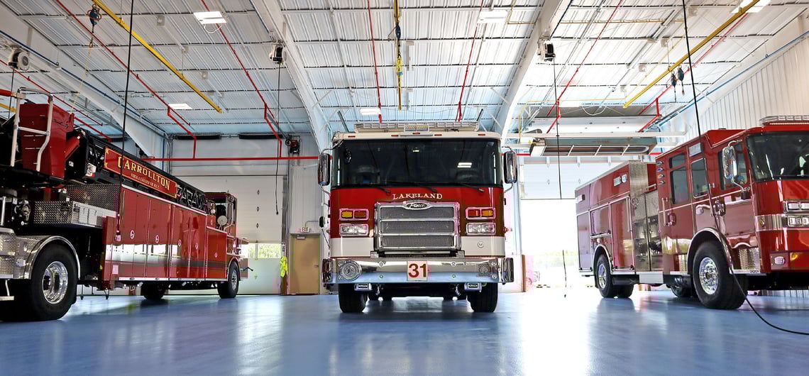 Three red fire trucks on the blue floor in a Pierce manufacturing building with one garage door open to the outside.