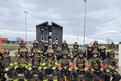 A group of firefighters in full turnout gear posing in front of a training tower.