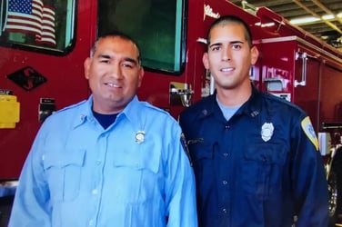 Two firefighters wearing blue uniforms stand in front of a red fire engine.