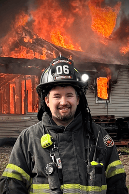 A smiling volunteer firefighter in protective gear with a house fire burning in the background.