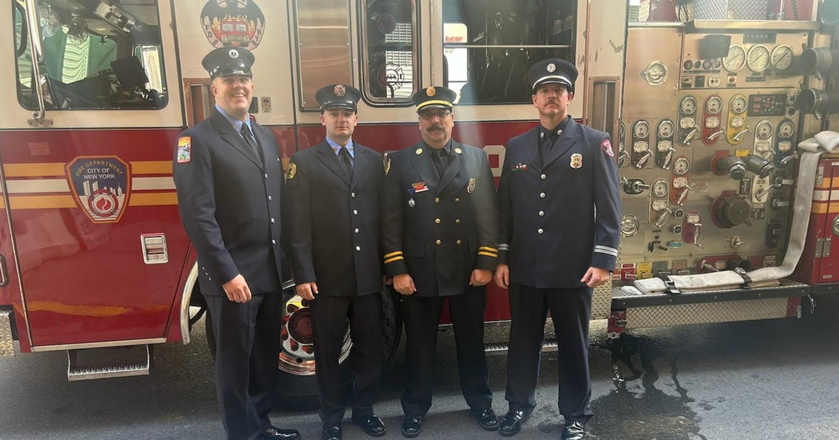Four FDNY firefighters in blue dress uniforms stand in front of a red fire engine.