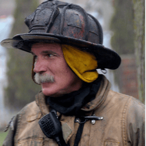 A side profile view of a firefighter in gear covered in soot and a helmet covered in soot as well.