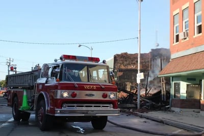 A red and white Pierce fire truck from Villa Grove is parked on a street with a damaged, smoking building in the background.