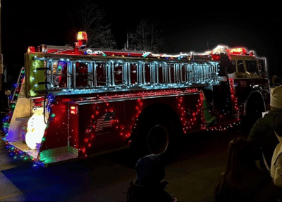 A vintage red fire truck, Engine 101, festively decorated with Christmas lights and a snowman, with Santa on board at a night parade.