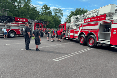 A group of people look at two red Pierce fire trucks, pareked on pavement.
