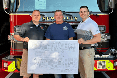Three people stand in front of a red fire truck, holding a custom-build document of their fire truck.