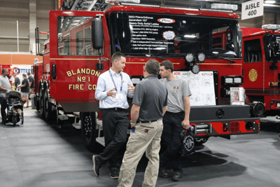 A salesperson talks to two people in front of a red fire truck on a tradeshow floor.