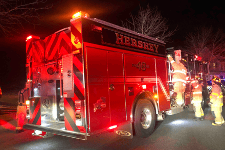 A red and black fire truck, readying Hershey at the top, has its lights on at night with three firefighters nearby.