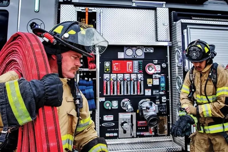Two firefighters in protective gear prepare equipment next to a Pierce fire truck.