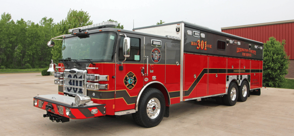 The red and gray Lexington County Fire Rescue service truck sits on the pavement with green trees and a red building in the background.