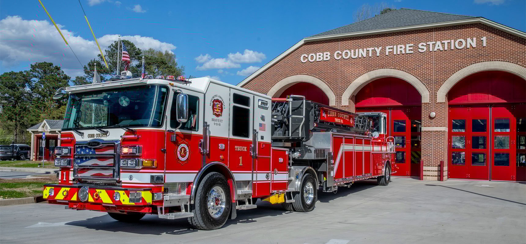 A red fire truck sitting out front of a brick fire station with the three red garage doors.