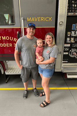 A family holding their young baby in front of a red and gray fire truck that reads 'Maverick'