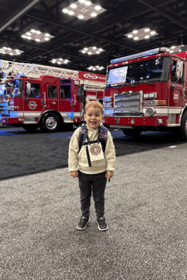 A young boy with a backpack and a badge on standing on a convention show floor with two red Pierce fire trucks in the background.