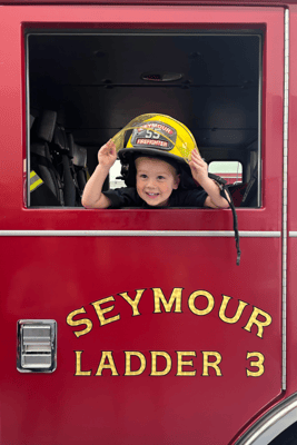 A young boy wearing a yellow fire fighters helmet inside a red fire truck that says Seymour Ladder 3 in gold letters on the side.