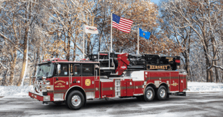 A red and black fire truck from Hershey, PA sitting on the pavement covered in snow with three flagpoles in the background.