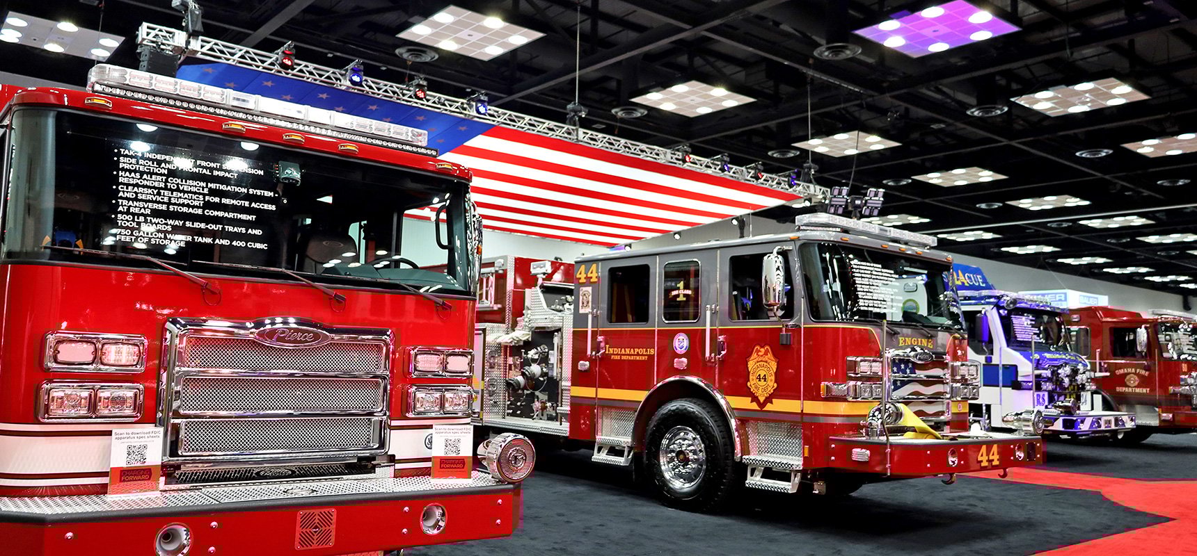 Two red fire trucks on a trade show floor with a large American flag hanging above.