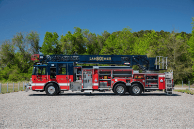 A red and black Pierce fire truck with a black aerial ladder sitting on a gravel road with green trees and blue sky in the background.